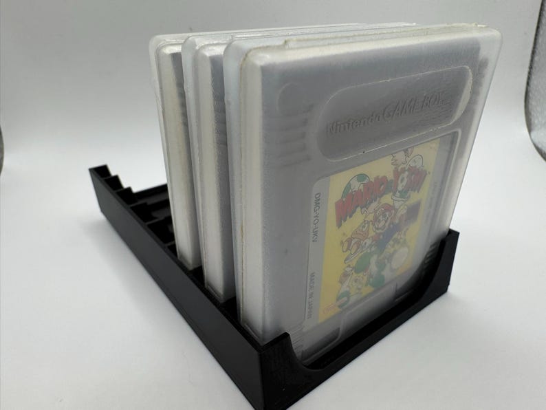 K&ouml;nnte beinhalten: Ein schwarzer Game-Cartridge-Halter aus Kunststoff mit vier transparenten Game-Cartridges aus Kunststoff. Die Game-Cartridges haben ein gelbes und farbenfrohes Etikett. Der Halter ist f&uuml;r mehrere Game-Cartridges konzipiert.