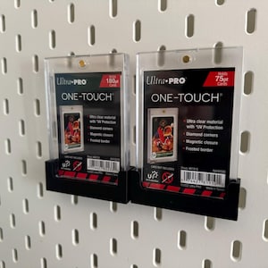 May include: Two clear Ultra Pro One-Touch card holders, one holding 180pt cards and the other 75pt cards. Each holder has a black base and features a card with a colorful image. The holders are mounted on a white pegboard.