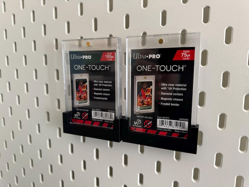 May include: Two clear Ultra Pro One-Touch card holders, one holding 180pt cards and the other 75pt cards. Each holder has a black base and features a card with a colorful image. The holders are mounted on a white pegboard.