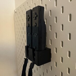 Puede incluir: Dos mandos de videojuegos negros est&aacute;n montados en una pared perforada blanca. Los mandos est&aacute;n en un soporte negro, con correas de mu&ntilde;eca negras colgando. La pared perforada tiene una serie de agujeros espaciados.