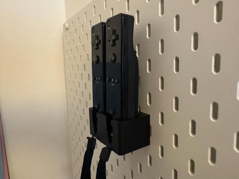 Puede incluir: Dos mandos de videojuegos negros est&aacute;n montados en una pared perforada blanca. Los mandos est&aacute;n en un soporte negro, con correas de mu&ntilde;eca negras colgando. La pared perforada tiene una serie de agujeros espaciados.