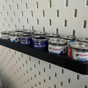 May include: A black shelf holds several small paint cans with silver lids. The cans are labeled with the brands "Revell Email Color" and "Humbrol Enamel." The shelf is mounted on a white pegboard.