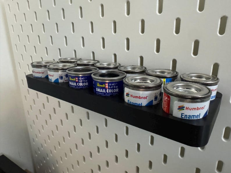 May include: A black shelf holds several small paint cans with silver lids. The cans are labeled with the brands "Revell Email Color" and "Humbrol Enamel." The shelf is mounted on a white pegboard.