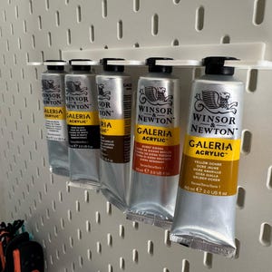May include: A selection of Winsor & Newton Galeria acrylic paint tubes, each holding 60 ml, arranged on a white pegboard. The tubes are silver with black caps and yellow labels, with various paint colours.