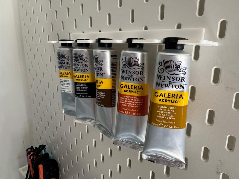 May include: A selection of Winsor & Newton Galeria acrylic paint tubes, each holding 60 ml, arranged on a white pegboard. The tubes are silver with black caps and yellow labels, with various paint colours.