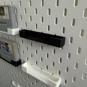 May include: A white pegboard with black and white shelves and game cartridges. The shelves are designed to hold items, and the cartridges are for a gaming console. The pegboard has evenly spaced oval holes.