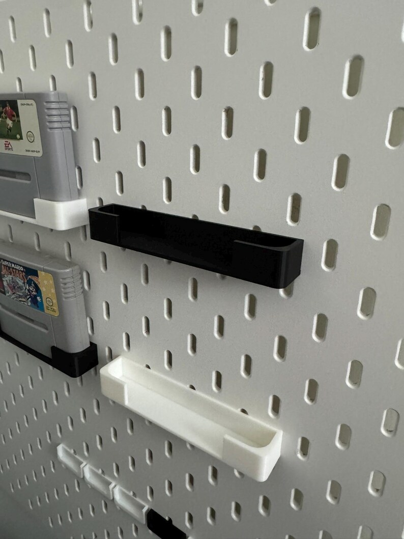 May include: A white pegboard with black and white shelves and game cartridges. The shelves are designed to hold items, and the cartridges are for a gaming console. The pegboard has evenly spaced oval holes.