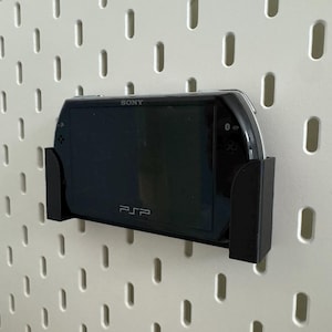 Pu&ograve; includere: Console di gioco portatile Sony PSP nera montata su una parete forata bianca con una staffa nera. La PSP mostra uno schermo scuro. Il logo PSP &egrave; visibile. La parete ha fori ovali.
