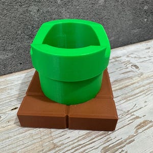 K&ouml;nnte beinhalten: Ein 3D-gedrucktes gr&uuml;nes Rohr, das an ein Videospiel erinnert, steht auf einer braunen rechteckigen Basis. Das Rohr hat eine abgerundete &Ouml;ffnung und einen zylindrischen K&ouml;rper. Die Basis besteht aus rechteckigen Bl&ouml;cken.