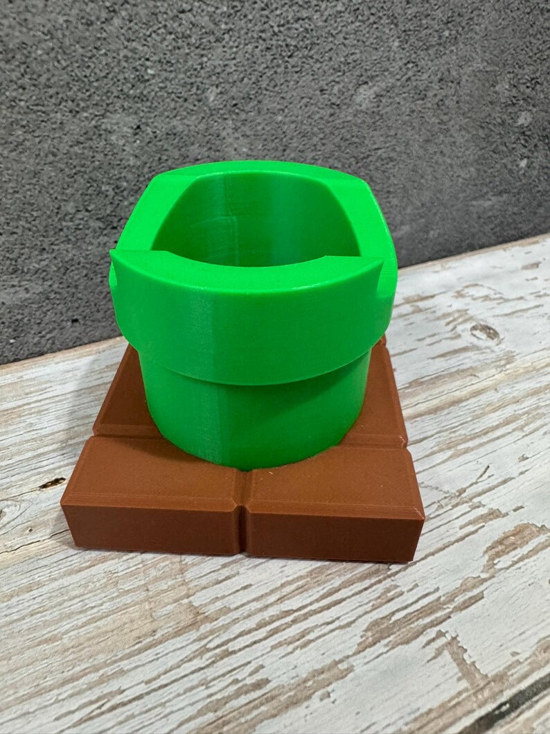 K&ouml;nnte beinhalten: Ein 3D-gedrucktes gr&uuml;nes Rohr, das an ein Videospiel erinnert, steht auf einer braunen rechteckigen Basis. Das Rohr hat eine abgerundete &Ouml;ffnung und einen zylindrischen K&ouml;rper. Die Basis besteht aus rechteckigen Bl&ouml;cken.