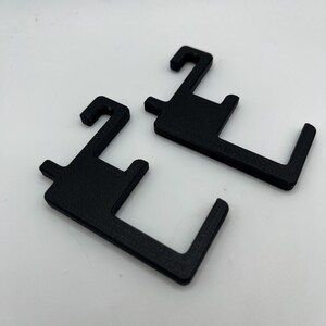 May include: Two black, 3D-printed plastic hooks. Each hook has a rectangular body with a curved hook at the top and a right-angled extension at the bottom. The hooks are designed for hanging items.