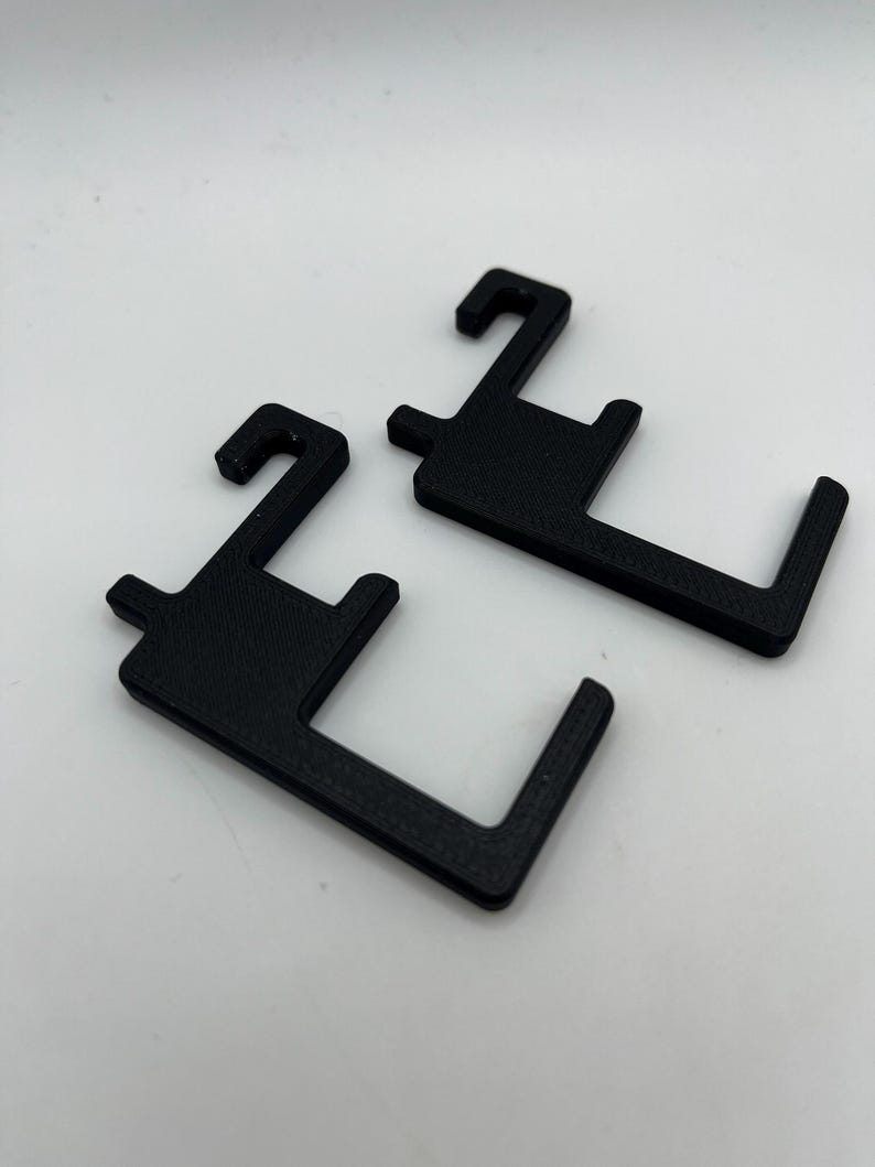 May include: Two black, 3D-printed plastic hooks. Each hook has a rectangular body with a curved hook at the top and a right-angled extension at the bottom. The hooks are designed for hanging items.