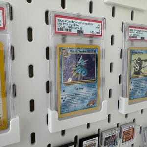 Peut inclure: Collection de cartes Pok&eacute;mon class&eacute;es, dont une carte Misty's Seadra, expos&eacute;es sur un panneau perfor&eacute; blanc. Les cartes sont dans des &eacute;tuis protecteurs avec des informations de classement. Les cartes sont dans des supports blancs.