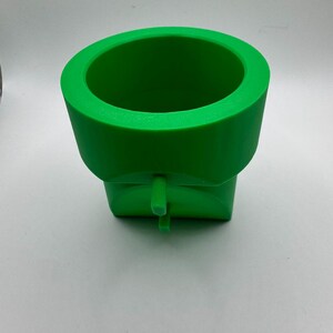 May include: A bright green, 3D-printed pipe, reminiscent of a video game. The cylindrical top has a wide opening, and the base is square. A small, rectangular tab extends from the front.