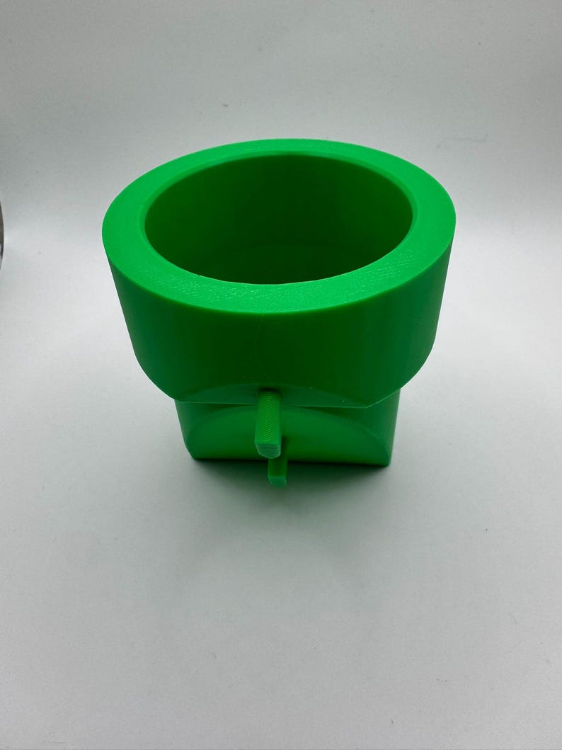 May include: A bright green, 3D-printed pipe, reminiscent of a video game. The cylindrical top has a wide opening, and the base is square. A small, rectangular tab extends from the front.