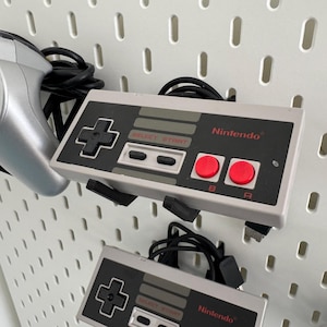 Puede incluir: Dos mandos grises de Nintendo Entertainment System con botones negros y botones A y B rojos. Los mandos est&aacute;n colgados en una pared blanca de tablero de clavijas.