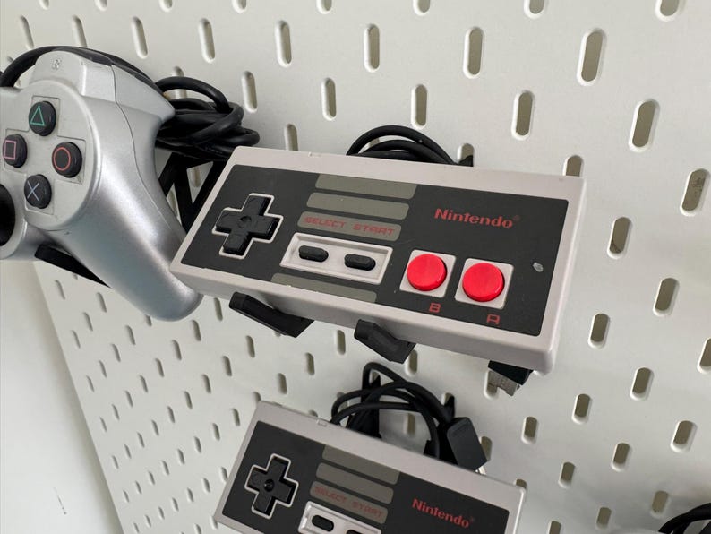 Puede incluir: Dos mandos grises de Nintendo Entertainment System con botones negros y botones A y B rojos. Los mandos est&aacute;n colgados en una pared blanca de tablero de clavijas.
