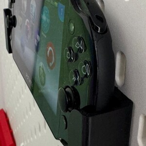 Op de afbeelding: Een zwarte PlayStation Vita handheld-gameconsole met een groen scherm is gemonteerd aan een witte pegboard-wand met behulp van een zwarte wandbeugel.