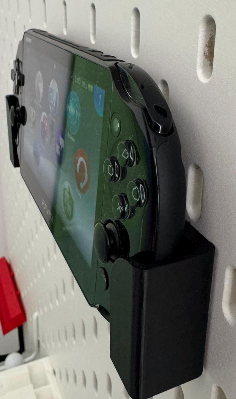 Op de afbeelding: Een zwarte PlayStation Vita handheld-gameconsole met een groen scherm is gemonteerd aan een witte pegboard-wand met behulp van een zwarte wandbeugel.