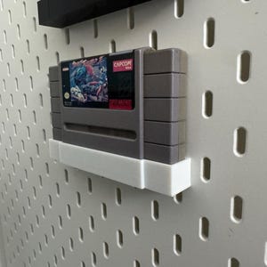 K&ouml;nnte beinhalten: Eine graue Super Nintendo-Spielkassette mit einem farbenfrohen Etikett mit Capcom-Logo und Artwork. Die Kassette ist auf einem wei&szlig;en Regal an einer wei&szlig;en Lochwand befestigt.