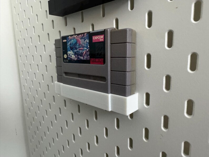 K&ouml;nnte beinhalten: Eine graue Super Nintendo-Spielkassette mit einem farbenfrohen Etikett mit Capcom-Logo und Artwork. Die Kassette ist auf einem wei&szlig;en Regal an einer wei&szlig;en Lochwand befestigt.
