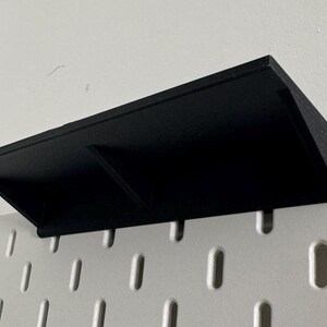 Puede incluir: Estante de pl&aacute;stico negro con dos compartimentos montado en una pared de tablero de clavijas blanco.