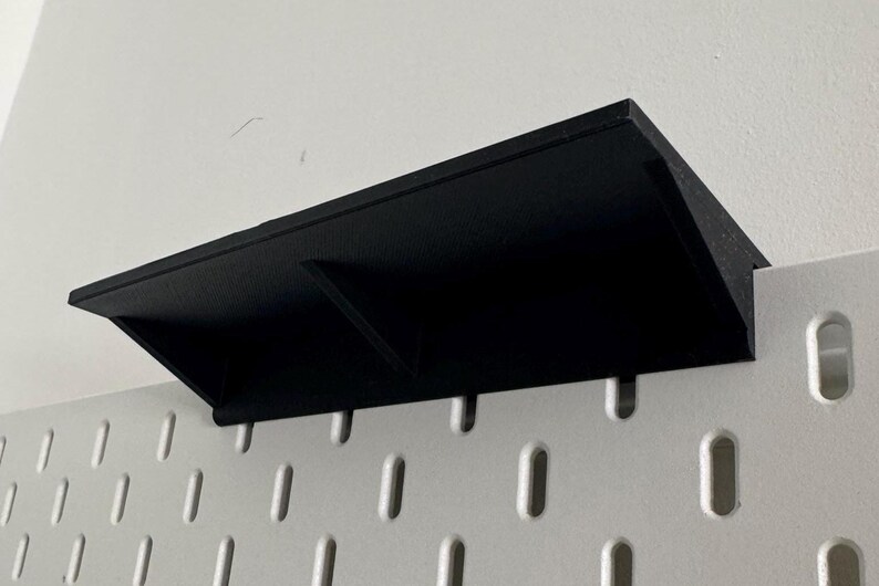 Puede incluir: Estante de pl&aacute;stico negro con dos compartimentos montado en una pared de tablero de clavijas blanco.