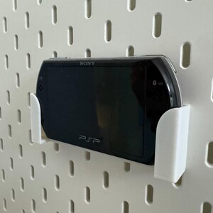 Pu&ograve; includere: Una console di gioco portatile Sony PSP nera &egrave; montata in un supporto bianco su una parete forata bianca. La PSP &egrave; tenuta saldamente dal supporto, mostrando lo schermo e il logo PSP.