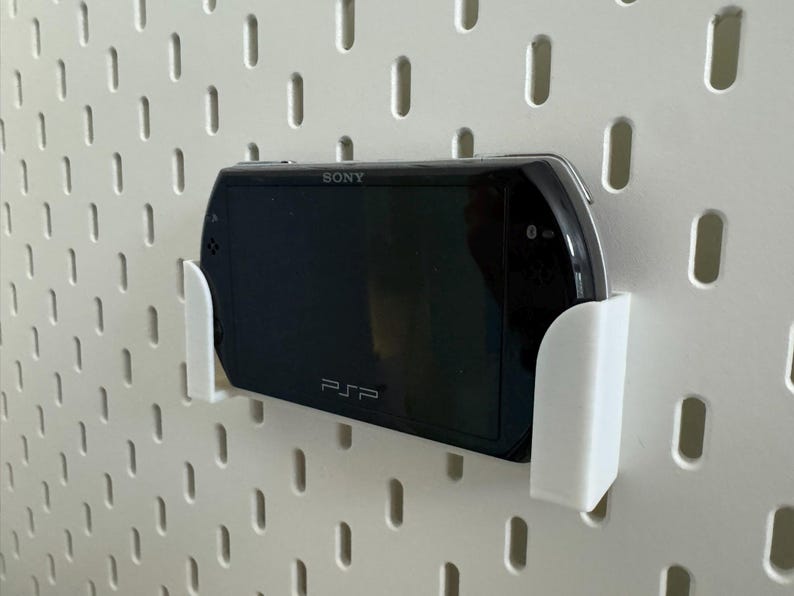 Pu&ograve; includere: Una console di gioco portatile Sony PSP nera &egrave; montata in un supporto bianco su una parete forata bianca. La PSP &egrave; tenuta saldamente dal supporto, mostrando lo schermo e il logo PSP.