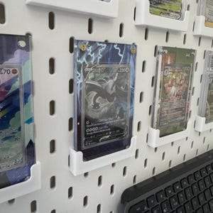 K&ouml;nnte beinhalten: Eine wei&szlig;e Lochwand mit mehreren durchsichtigen Plastikkartenhaltern, die verschiedene Pok&eacute;mon-Sammelkarten zeigen. Die Karten sind in Reihen und Spalten angeordnet, wobei einige Karten holografische Designs aufweisen. Eine schwarze Tastatur ist teilweise in der unteren rechten Ecke des Bildes zu sehen.