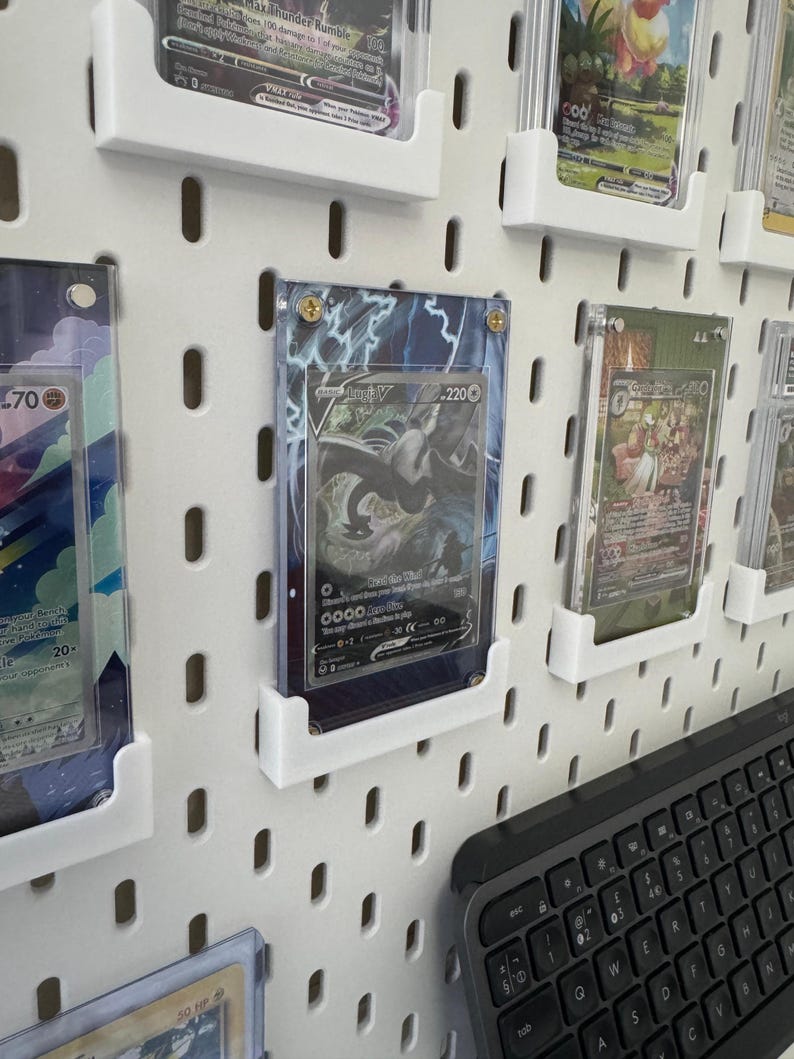 K&ouml;nnte beinhalten: Eine wei&szlig;e Lochwand mit mehreren durchsichtigen Plastikkartenhaltern, die verschiedene Pok&eacute;mon-Sammelkarten zeigen. Die Karten sind in Reihen und Spalten angeordnet, wobei einige Karten holografische Designs aufweisen. Eine schwarze Tastatur ist teilweise in der unteren rechten Ecke des Bildes zu sehen.