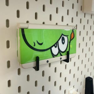 以下が含まれることがあります： 白いペグボードに取り付けられた、漫画の目のデザインが施されたライムグリーンの携帯ゲーム機。コンソールは2つの黒いブラケットで固定されています。ペグボードには、等間隔に配置された楕円形の穴があります。