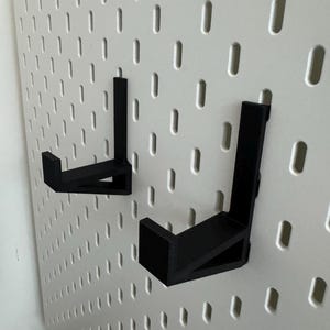 May include: Two black, L-shaped hooks are mounted on a white pegboard. The hooks are designed to hold items and are made of a solid, matte material. The pegboard has evenly spaced holes.