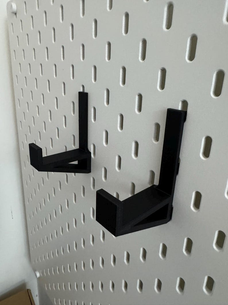 May include: Two black, L-shaped hooks are mounted on a white pegboard. The hooks are designed to hold items and are made of a solid, matte material. The pegboard has evenly spaced holes.