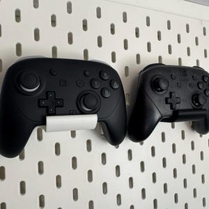 K&ouml;nnte beinhalten: Zwei schwarze Videospiel-Controller, einer gr&ouml;&szlig;er als der andere, an einer wei&szlig;en Lochwand befestigt. Der gr&ouml;&szlig;ere Controller hat eine wei&szlig;e Halterung, w&auml;hrend der kleinere eine schwarze Halterung hat. Die Controller sind zum Spielen.