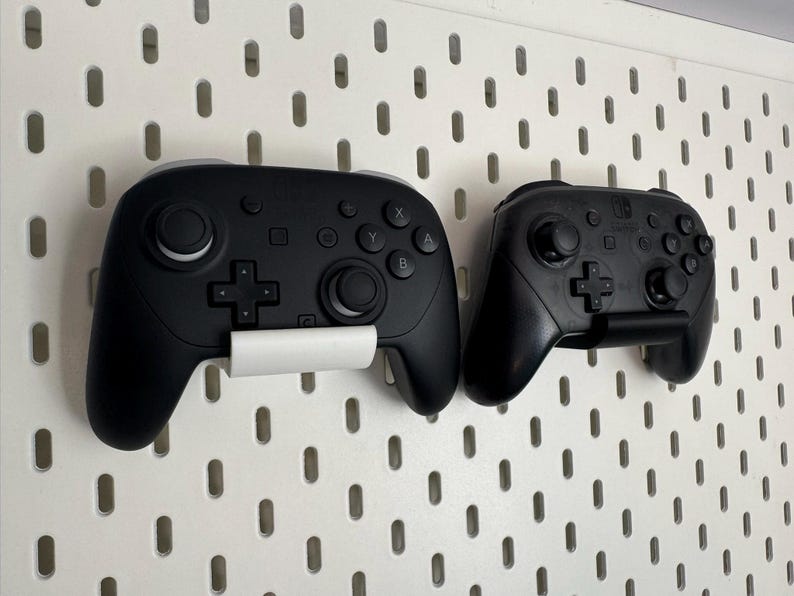 K&ouml;nnte beinhalten: Zwei schwarze Videospiel-Controller, einer gr&ouml;&szlig;er als der andere, an einer wei&szlig;en Lochwand befestigt. Der gr&ouml;&szlig;ere Controller hat eine wei&szlig;e Halterung, w&auml;hrend der kleinere eine schwarze Halterung hat. Die Controller sind zum Spielen.