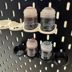 Puede incluir: Un soporte de pl&aacute;stico blanco para botes de pintura con cuatro ranuras, que contiene cuatro botes de pintura Citadel Colour. Los botes est&aacute;n etiquetados con "Citadel Colour" y tienen diferentes tonos de pintura marr&oacute;n y negro.