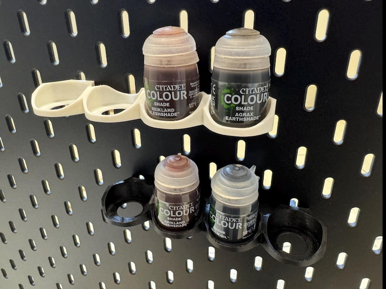 Puede incluir: Un soporte de pl&aacute;stico blanco para botes de pintura con cuatro ranuras, que contiene cuatro botes de pintura Citadel Colour. Los botes est&aacute;n etiquetados con "Citadel Colour" y tienen diferentes tonos de pintura marr&oacute;n y negro.