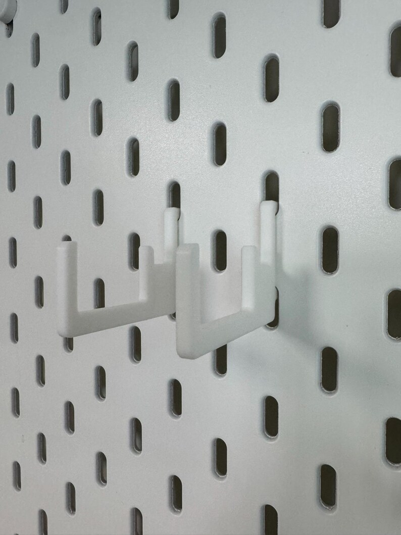 May include: Two white pegboard hooks are inserted into a white pegboard. The hooks are designed to hold items and are made of a durable plastic material. The pegboard has evenly spaced oval holes.