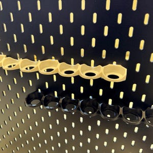 以下が含まれることがあります： 黒いペグボードの壁に、ベージュのプラスチック製のペグボードフックが2列取り付けられています。各列には7つのフックがあります。