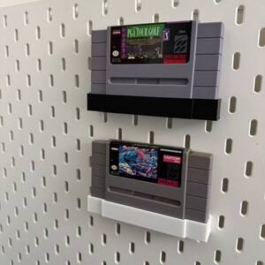 K&ouml;nnte beinhalten: Zwei Super Nintendo-Spielmodule, die auf einer wei&szlig;en Lochwand ausgestellt sind. Das obere Modul ist "PGA Tour Golf" und das untere ist "Street Fighter 2". Beide sind grau mit farbenfrohen Etiketten und schwarzen oder wei&szlig;en Haltern.