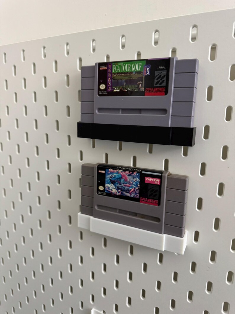 K&ouml;nnte beinhalten: Zwei Super Nintendo-Spielmodule, die auf einer wei&szlig;en Lochwand ausgestellt sind. Das obere Modul ist "PGA Tour Golf" und das untere ist "Street Fighter 2". Beide sind grau mit farbenfrohen Etiketten und schwarzen oder wei&szlig;en Haltern.