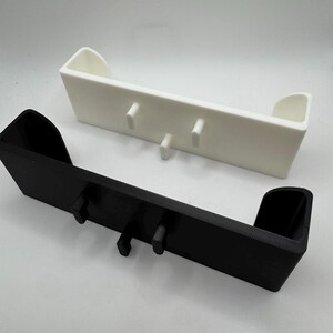 Puede incluir: Dos estantes de pared impresos en 3D. Uno es negro, el otro blanco. Cada estante tiene un dise&ntilde;o rectangular con dos clavijas verticales para colgar objetos. Los estantes tienen un peque&ntilde;o labio en cada extremo.