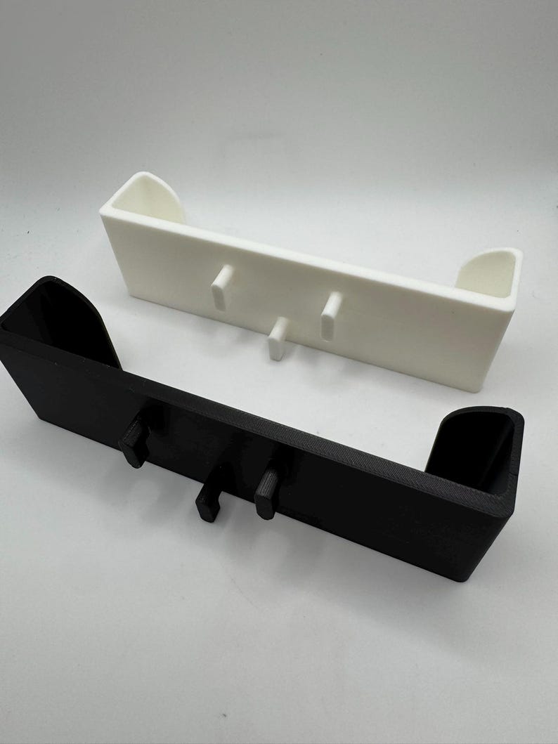 Puede incluir: Dos estantes de pared impresos en 3D. Uno es negro, el otro blanco. Cada estante tiene un dise&ntilde;o rectangular con dos clavijas verticales para colgar objetos. Los estantes tienen un peque&ntilde;o labio en cada extremo.