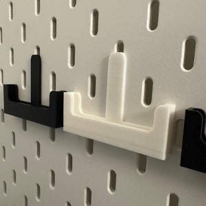 Op de afbeelding: Close-up van zwarte en witte pegboard haken. De haken hebben de vorm van een "U" met een verticale balk. De haken zijn in een wit pegboard met gelijkmatig verdeelde gaten geplaatst.