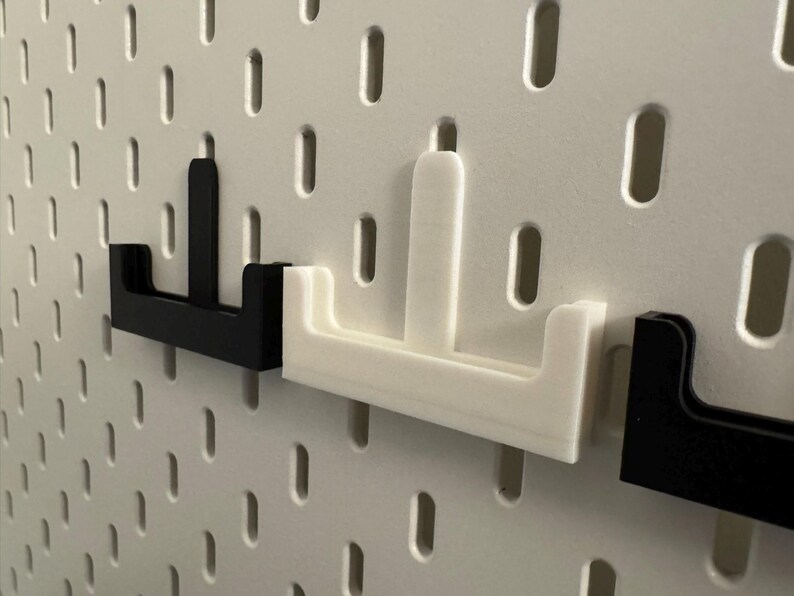 Op de afbeelding: Close-up van zwarte en witte pegboard haken. De haken hebben de vorm van een "U" met een verticale balk. De haken zijn in een wit pegboard met gelijkmatig verdeelde gaten geplaatst.