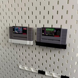 K&ouml;nnte beinhalten: Zwei Retro-Videospielkassetten, eine mit wei&szlig;em und die andere mit schwarzem Sockel, werden auf einer wei&szlig;en Lochwand ausgestellt. Die Kassetten sind grau mit farbenfrohen Etiketten. Ein Spiel tr&auml;gt die Aufschrift "PGA TOUR GOLF".