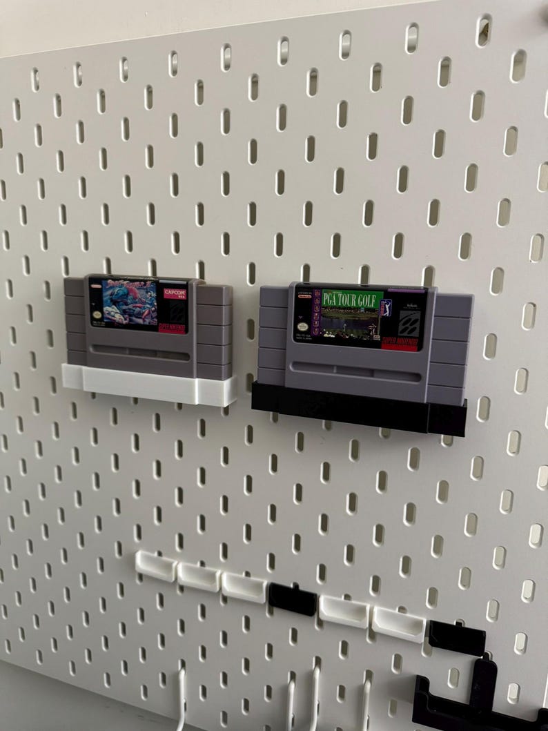 K&ouml;nnte beinhalten: Zwei Retro-Videospielkassetten, eine mit wei&szlig;em und die andere mit schwarzem Sockel, werden auf einer wei&szlig;en Lochwand ausgestellt. Die Kassetten sind grau mit farbenfrohen Etiketten. Ein Spiel tr&auml;gt die Aufschrift "PGA TOUR GOLF".