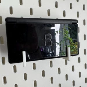 Puede incluir: Una consola de juegos port&aacute;til Nintendo DS Lite negra, exhibida en una pared perforada blanca con soportes blancos. La pantalla refleja una planta verde. La consola tiene dos pantallas y un conjunto de botones de control.