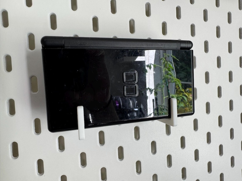 Puede incluir: Una consola de juegos port&aacute;til Nintendo DS Lite negra, exhibida en una pared perforada blanca con soportes blancos. La pantalla refleja una planta verde. La consola tiene dos pantallas y un conjunto de botones de control.
